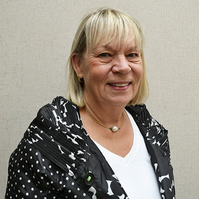 A smiling woman with short blonde hair wearing a black and white patterned jacket and a white shirt against a neutral background.