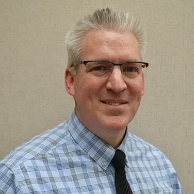 A smiling man with silver hair wearing glasses and a checked shirt with a tie.